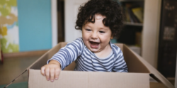 A young child with curly hair, wearing a striped shirt, sits inside a cardboard box, smiling and reaching forward excitedly.