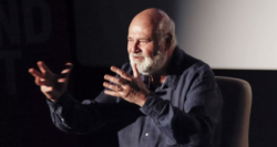 An older man with a white beard and dark shirt sits in a chair, gesturing expressively with both hands, in front of a dark background and a projected screen.