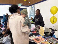 A woman holds a baby while speaking to a woman at a table with books, handouts, and yellow balloons at an indoor community event.