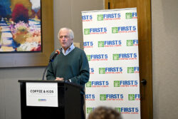 An older man speaks at a podium labeled Coffee & Kids, with a First 5 Contra Costa banner in the background. He is wearing a green sweater and a collared shirt.