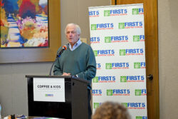 An older man speaks at a podium labeled Coffee & Kids with a microphone. Behind him is a banner with the First 5 Contra Costa logo. A colorful painting is on the wall to his left.