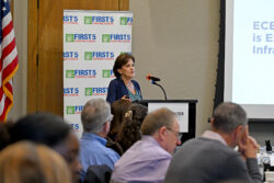 A woman speaks at a podium during a conference. The audience listens as she stands beside a screen and a First 5 Contra Costa banner. An American flag is visible in the background.