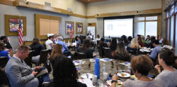 People attend a conference in a large room. A speaker presents at the front near a screen. Attendees sit at round tables with food, drinks, and papers. American flags are displayed in the room.