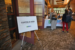 A sign reading Coffee & Kids stands near an entrance, while people gather and check in at a registration table inside a well-lit event space.
