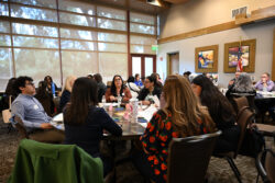 A group of people sit around tables in a conference room, engaged in conversation and activities. Large windows let in light, and colorful art hangs on the walls.