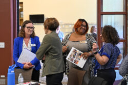Four women stand and talk around a conference table. Two hold documents, one with a collage of photos. Bottles and papers are on the table, and a glass wall is in the background. It appears to be a professional meeting.