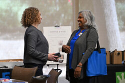 Two women stand and talk, smiling at each other during an event. One holds a phone, and the other carries a blue tote bag. A Coffee & Kids event sign is visible in the background.