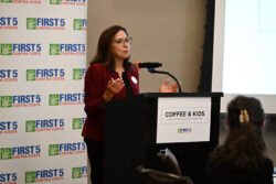 A woman in a red blazer speaks at a podium labeled COFFEE & KIDS during a First 5 Contra Costa event, with an audience and a branded backdrop behind her.