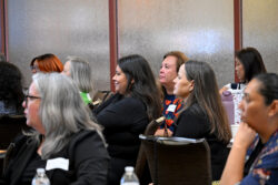 Several women sit at tables, attentively listening during an indoor event. They face forward, and some have name tags. Water bottles and cups are visible on the tables. The background shows frosted glass panels.
