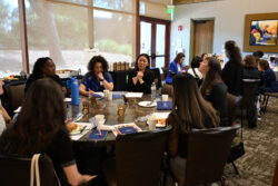 A group of women sit around a table in a conference room, engaged in discussion with notebooks, coffee cups, and snacks in front of them. Other small groups are visible in the background.
