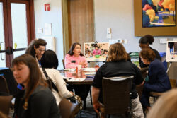 People sit around tables in a conference room, engaged in conversation. A woman in pink speaks at one table. Posters, coffee cups, and paperwork are visible. Paintings hang on the wall in the background.