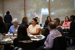 People sit at round tables engaged in conversation during a meeting or workshop. Papers, drinks, and snacks are on the tables. Large windows in the background let in natural light.