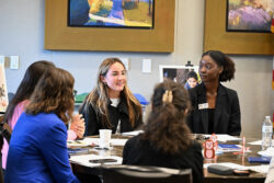 Five women sit around a table in discussion, with papers and drinks in front of them. Two women face the camera, smiling, while three others have their backs to the camera. Colorful artwork hangs on the wall behind them.