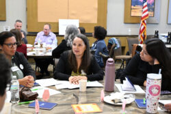 Four women sit around a table in discussion, surrounded by papers, notebooks, and drinks; other groups and an American flag are visible in the background at a professional meeting.