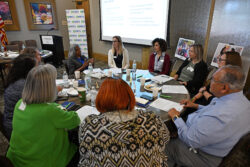 A diverse group of people sit around a table in discussion, with papers, laptops, and drinks. A presentation is displayed on a screen, and photos are visible in the background.