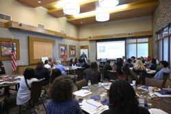 A group of people sit at round tables in a conference room, watching a speaker present a slideshow at the front. The room has large windows, wall art, and an American flag.