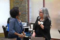 Two women stand indoors having a conversation. One woman has curly hair, wears a blue outfit, and holds a cup; the other has gray hair, glasses, and wears black. There are chairs and tables in the background.