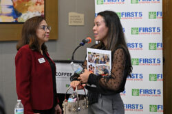 Two women talk at a First 5 Contra Costa event; one holds a folder with photos and a water bottle, the other stands near a podium. A First 5 Contra Costa step and repeat banner is in the background.