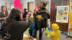 A man holding a young child smiles while showing a t-shirt. A woman takes his photo, and a child in a yellow raincoat with balloon animals stands nearby. Other people are in the background at an indoor event.