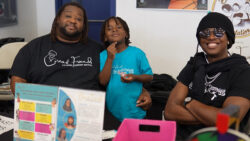 Three people sit at a table covered with pamphlets and colorful items. One adult smiles, another wears sunglasses and crosses arms, and a child in the middle gives a thumbs-up, all wearing matching event shirts.