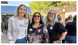 Three smiling women stand together outdoors at an event, wearing name tags and buttons. Other people and event signs are visible in the background. The setting appears casual and friendly.
