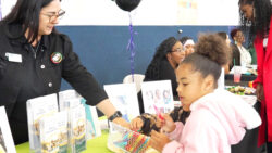 A woman stands behind a table with brochures, handing candy to a young girl in a pink jacket. Other children and adults are gathered around the table at what appears to be a community event.