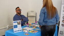 A man sits at a blue table with pamphlets, bubbles, and toys, talking to a woman standing across from him. Both are indoors near a cream wall and white chair.