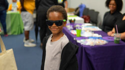 A young boy wearing sunglasses and a black jacket smiles at the camera in a room with tables covered in purple and green cloths, with people in the background.