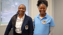 Two women stand indoors, smiling at the camera. Both wear uniforms with matching embroidered badges. One wears a navy blazer over a white shirt, and the other wears a blue polo shirt over a striped long-sleeve top.