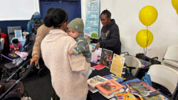 A woman holding a baby speaks to a vendor at a table covered with colorful childrens books, pamphlets, and yellow balloons at an indoor event. Other attendees are visible in the background.