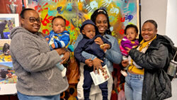 Three women stand smiling, each holding a young child, in front of a colorful handprint mural. Another woman stands at the end, smiling. The scene appears cheerful and lively.