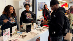 A group of people interact at an indoor event booth with brochures and informational posters on the table; one person looks at their phone while others stand nearby, smiling and talking.