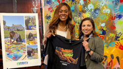 Two women smile, holding a black T-shirt with colorful text. Beside them is a poster with photos and the FIRST 5 Contra Costa logo, showing children and adults creating handprint art outdoors. Colorful handprints cover the wall behind.