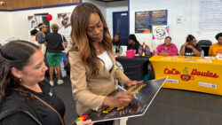 A woman in a tan suit signs a poster while another woman watches at an indoor event with informational booths, people seated at tables, and colorful displays in the background.