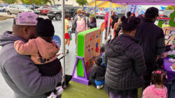 Adults and children gather under a tent at an outdoor event, exploring colorful activity boards and books. Some adults hold young children, and kids wear backpacks. Cars and mural-painted walls are visible in the background.
