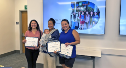 Three women stand and smile holding certificates; one also holds a baby. They are in a classroom with a large screen behind them showing a Thank You slide and a group photo.