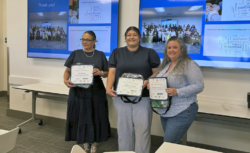 Three women stand indoors, smiling and holding certificates and bags. Behind them, three large screens display group photos, a word cloud, and Thank you! in a classroom or conference setting.