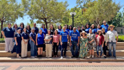 A group of about 30 people, some wearing blue Grandma & Me shirts, pose together outside on steps, surrounded by trees and sunlight.