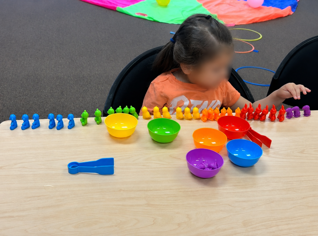 A child sits at a table, sorting colorful bear-shaped counters into matching bowls. The bears and bowls are arranged by color: blue, green, yellow, orange, red, and purple. The childs face is blurred for privacy.