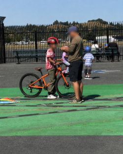 A child wearing a helmet stands with an orange bike next to an adult on a painted area, with people sitting on benches and children playing in the background.
