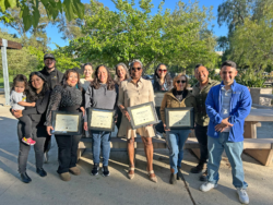A group of people stand outdoors, some holding certificates and smiling at the camera. There are trees and picnic tables in the background, and the setting is bright and sunny.