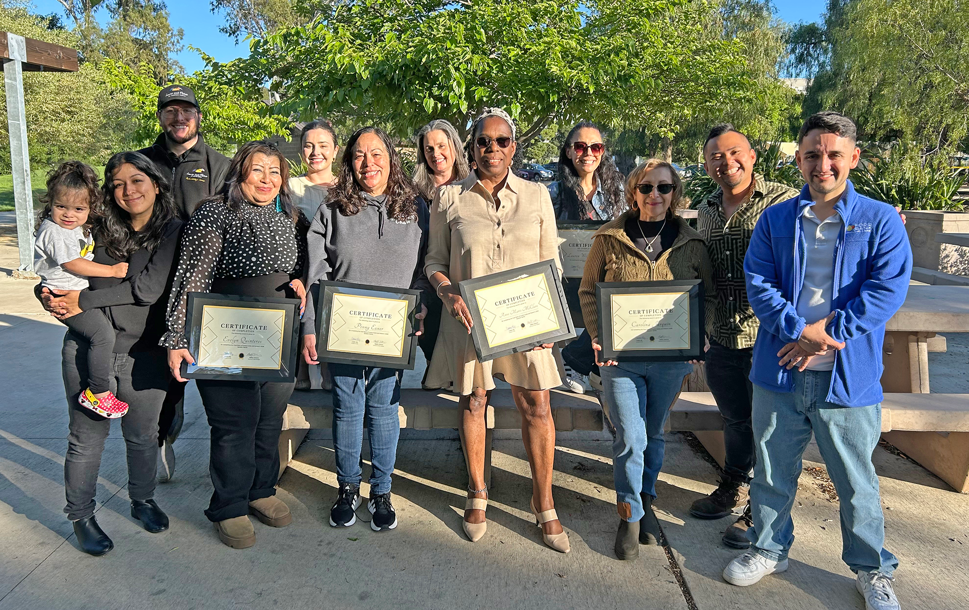 A group of people poses outdoors, some holding certificates and smiling. There are trees and a picnic table in the background. One person holds a small child on the left side of the group.