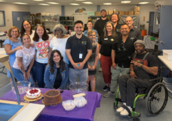 A group of people smiling and posing together in a kitchen, with cakes and desserts on a table in front of them. One person is in a wheelchair. The setting appears to be cheerful and communal.