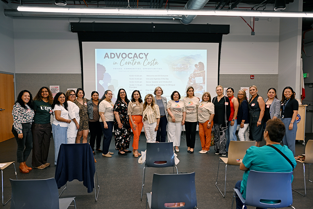 A group of people stand in front of a screen displaying Advocacy in Contra Costa in a classroom or conference room, with empty chairs in the foreground.