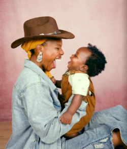 A woman in a hat and denim jacket holds a smiling baby wearing brown overalls. Both are laughing and facing each other against a pink background.