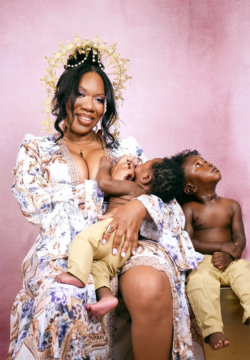 A woman in a patterned dress wears a gold crown and breastfeeds a baby. Another young child sits beside her, gazing upward. The background is pink.