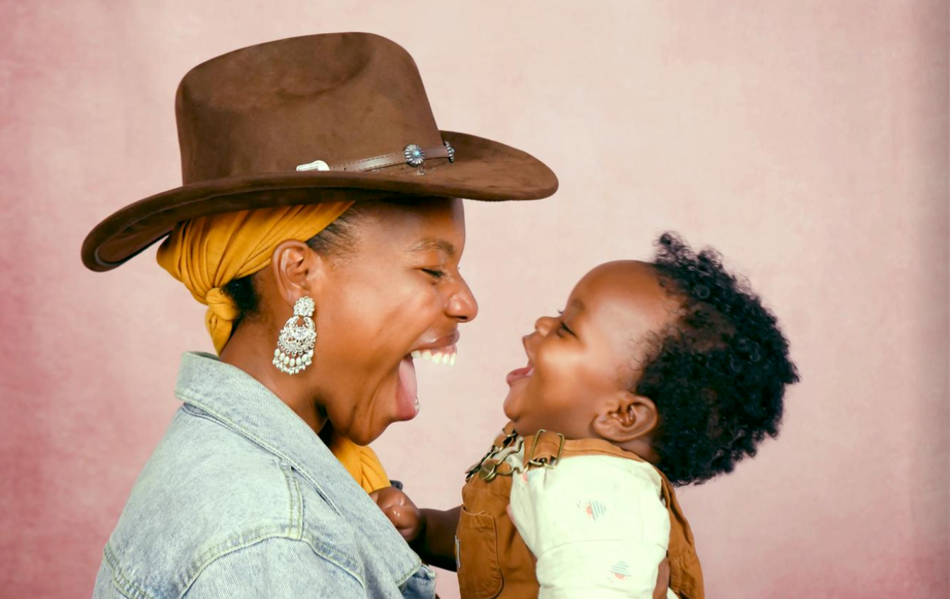 A woman in a cowboy hat and earrings holds a laughing baby, both smiling widely at each other, in front of a pink background.