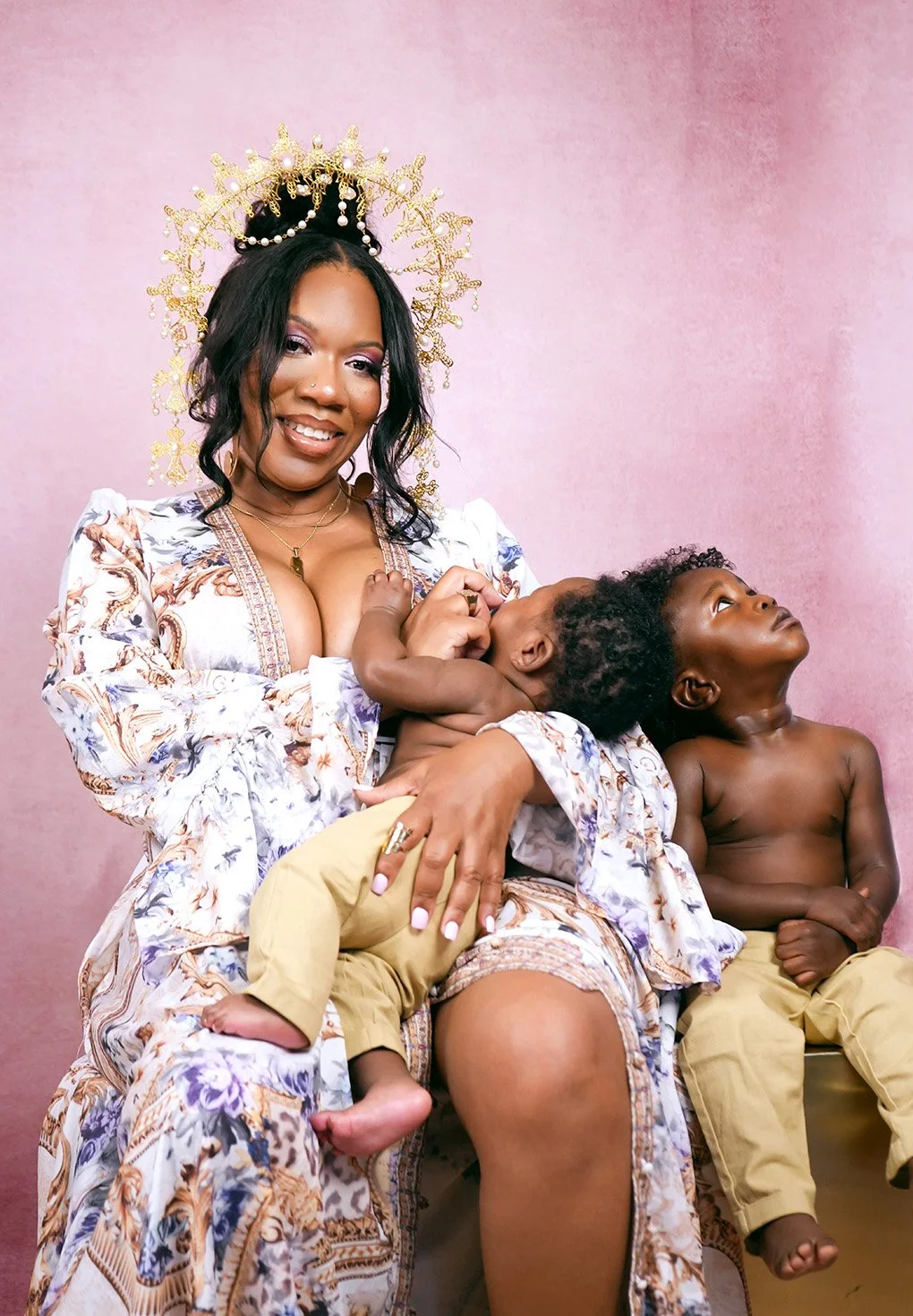 A woman in a patterned dress wears a gold crown and breastfeeds a baby. Another young child sits beside her, gazing upward. The background is pink.