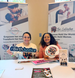 Two women sit at a table with flyers, smiling, holding #BirthJustice and Black girls mental health signs. Behind them are banners promoting perinatal mental health and Black Girls Mental Health Collective.