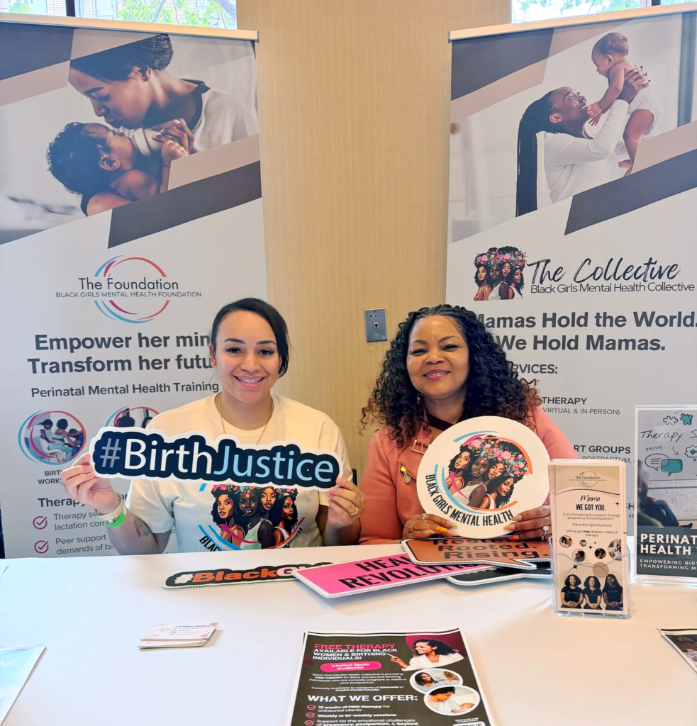 Two women sit at a table with flyers, smiling, holding #BirthJustice and Black girls mental health signs. Behind them are banners promoting perinatal mental health and Black Girls Mental Health Collective.